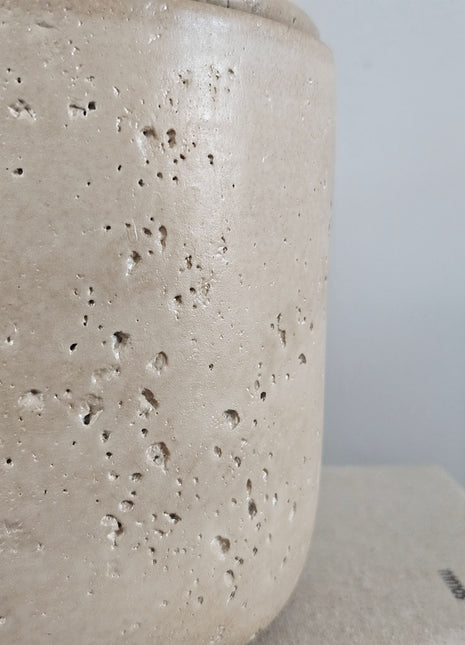 Close-up van een beige potje met deksel met een gespikkelde structuur en een rond knopje op een lichte achtergrond