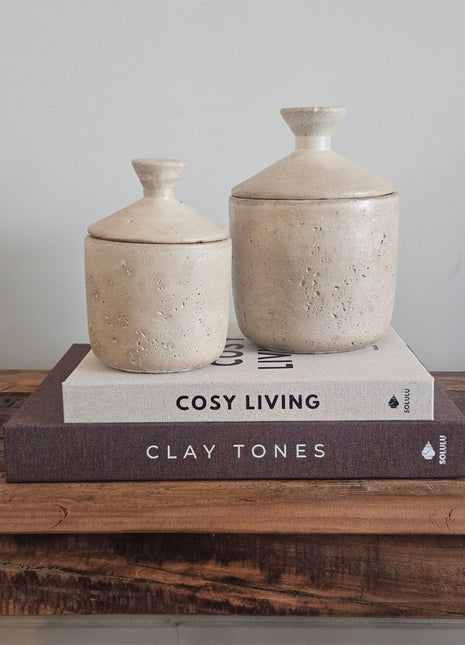 Set van twee aardewerk potjes met deksel beige in maat S en M op koffietafelboeken Cosy Living en Clay Tones op een houten ondergrond