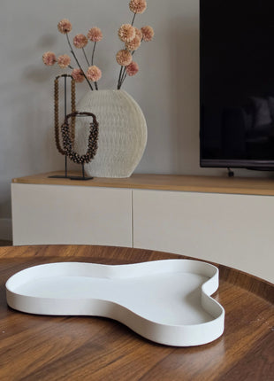 Dienblad Amy Ivory in druppelvorm van wit mat keramiek op een houten tafel naast twee amber glazen kandelaars met witte dinerkaarsen en een witte bolbloem