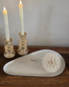 Dienblad Nova Offwhite in druppelvorm van off-white mat keramiek op een houten tafel naast twee amber glazen kandelaars met witte dinerkaarsen en een witte paardenbloem