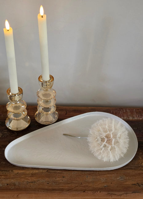 Dienblad Nova Offwhite in druppelvorm van off-white mat keramiek op een houten tafel naast twee amber glazen kandelaars met witte dinerkaarsen en een witte paardenbloem