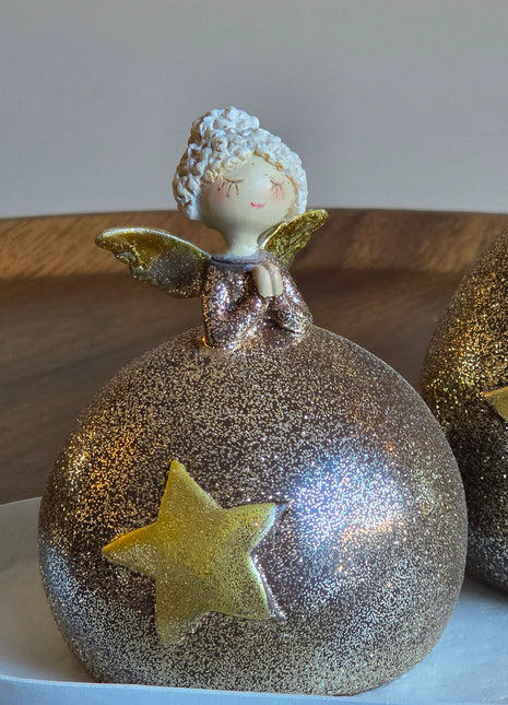 Close-up van engeltje S met witte krullende haren en gouden vleugels op een ronde glitterbol in goud-brons met een gouden ster op een wit oppervlak