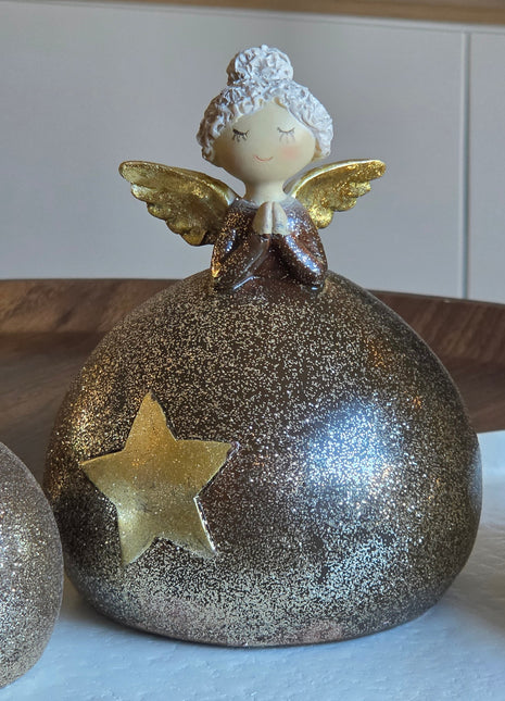 Close-up van engeltje M met witte krullende haren en gouden vleugels op een ronde glitterbol in goud-brons met een gouden ster op een houten ondergrond