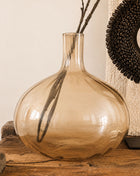 Glazen vaas bruin amber glanzend L met bolle ronde vorm en smalle hals op een houten dressoir met droge takken en een donkere schelpenketting op een standaard