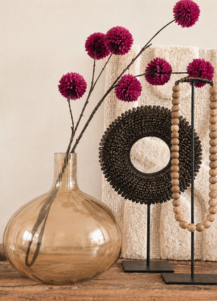 Glazen vaas bruin amber glanzend L met bordeauxrode pompom zijdetakken naast een donkere schelpenketting en een houten kralenketting op een standaard voor een crème wandpaneel