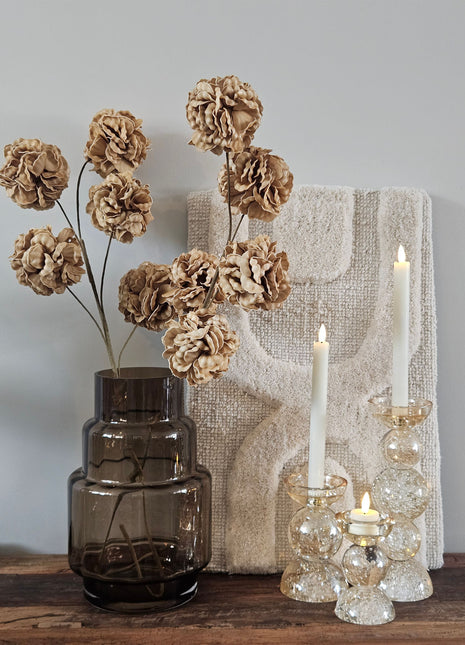 Cilindrisch taupe rookglazen windlicht Dune Taupe S met een brandende LED-kaars op een rond bronzen bijzettafeltje voor een lichte muur