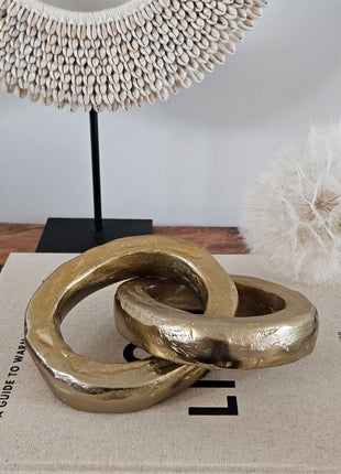 Close-up van twee ineen gehaakte gouden ringen van aluminium met ruwe gegoten structuur op een wit koffietafelboek naast een witte schelpenketting op standaard