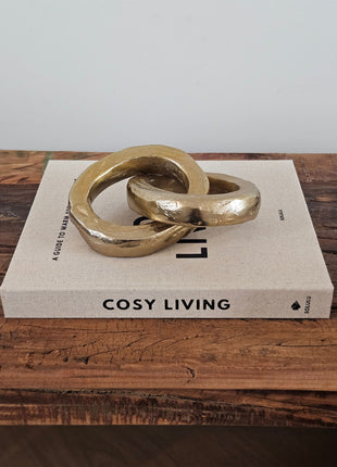 Twee ineen gehaakte gouden ringen van aluminium op een wit koffietafelboek "Cosy Living" op een houten ondergrond