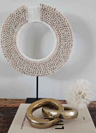 Twee ineen gehaakte gouden ringen van aluminium op een wit koffietafelboek naast een witte schelpenketting op zwarte standaard en een witte gedroogde paardenbloem op een houten ondergrond