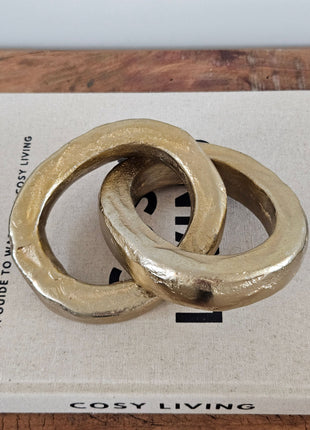 Twee ineen gehaakte gouden ringen van aluminium met ruwe gegoten structuur op een wit koffietafelboek op een houten ondergrond