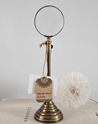 Gouden vergrootglas op messing standaard met ronde voet op een houten ondergrond naast een witte vaas