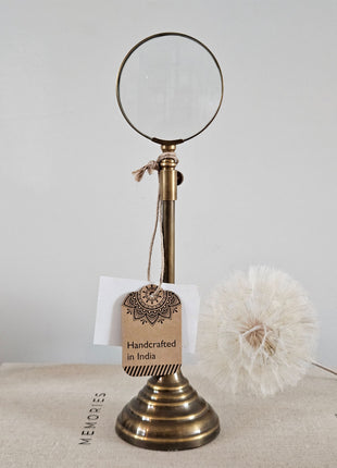 Gouden vergrootglas op messing standaard met ronde voet op een houten ondergrond naast een witte vaas