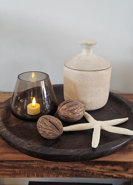 Rond donker houten dienblad met twee kleine handvatten met een rookglazen windlicht met LED-kaars  een beige potje met deksel  twee droge noten en een witte zeester op een houten ondergrond