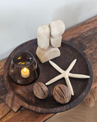 Rond donker houten dienblad met twee kleine handvatten met een rookglazen windlicht met LED-kaars  een beige ornament  een witte zeester en twee droge noten op een houten ondergrond