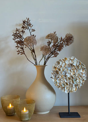 Vooraanzicht van goudkleurige Eupatorium kunsttakken met glanzende bloemschermen in creme vaas naast schelpen op hout