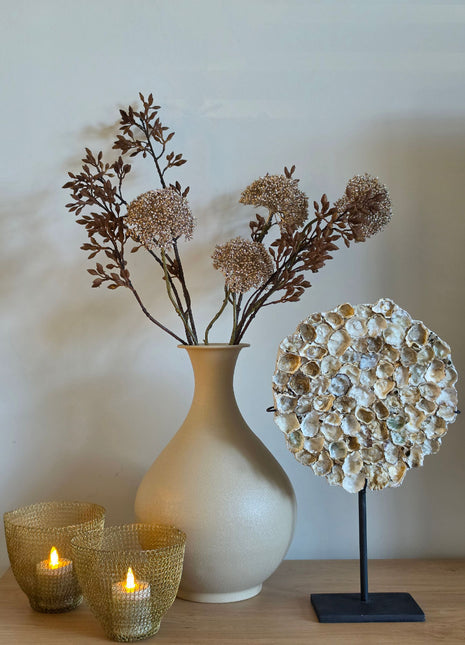 Vooraanzicht van goudkleurige Eupatorium kunsttakken met glanzende bloemschermen in creme vaas naast schelpen op hout