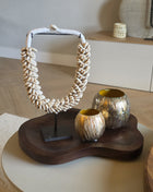 Een decoratieve interieurset op een organisch gevormd, donkerhouten dienblad met een grote schelpenketting van kauri-schelpen op een zwarte standaard en twee ronde, metallic goudkleurige windlichten met textuur.