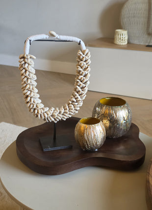 Een decoratieve interieurset op een organisch gevormd, donkerhouten dienblad met een grote schelpenketting van kauri-schelpen op een zwarte standaard en twee ronde, metallic goudkleurige windlichten met textuur.
