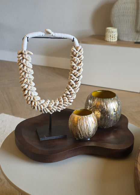 Een decoratieve interieurset op een organisch gevormd, donkerhouten dienblad met een grote schelpenketting van kauri-schelpen op een zwarte standaard en twee ronde, metallic goudkleurige windlichten met textuur.