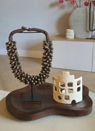 Een sfeervolle interieurset gepresenteerd op een organisch gevormd, donkerhouten dienblad met een bruine schelpenketting op een zwarte standaard en een crèmewit windlicht met uitgesneden raampjes in de vorm van een huisje.