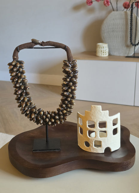 Een sfeervolle interieurset gepresenteerd op een organisch gevormd, donkerhouten dienblad met een bruine schelpenketting op een zwarte standaard en een crèmewit windlicht met uitgesneden raampjes in de vorm van een huisje.