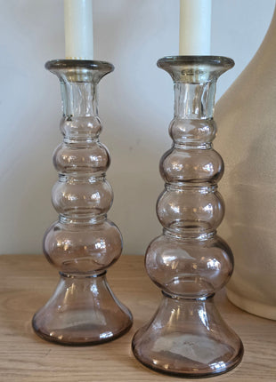 Close-up van twee rookbruine Jade glazen kandelaars met ronde bollen en witte kaarsen naast een creme vaas op hout