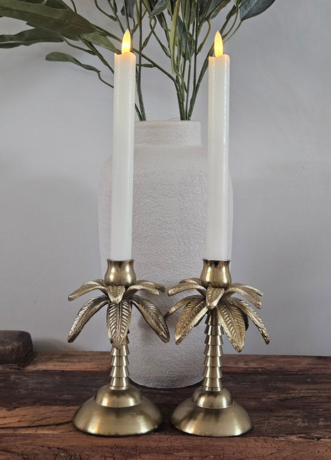 Kandelaar palmboom goud aluminium met een witte kaars naast een schelpenketting op een zwarte metalen standaard op een houten ondergrond