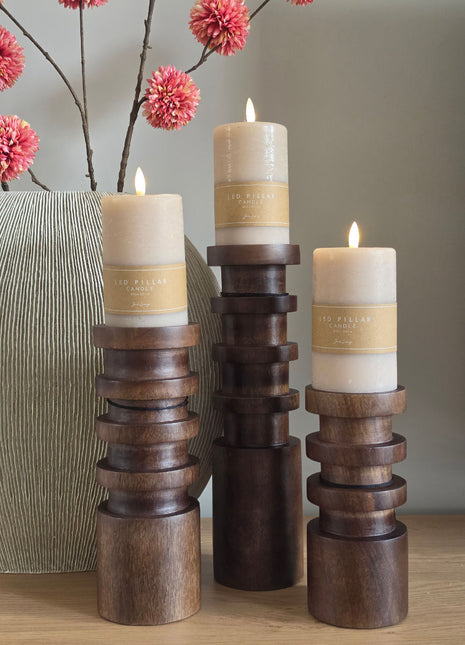 Vooraanzicht van drie Vail kandelaars in bruin hout met witte LED kaarsen naast roze bloemen op een houten tafel