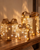 Vooraanzicht van drie goudkleurige kerstcadeautjes van metaaldraad met strikken en LED lichtjes op een houten bank