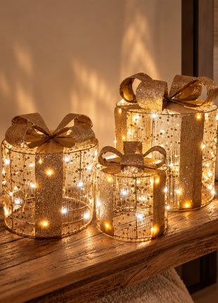 Vooraanzicht van drie goudkleurige kerstcadeautjes van metaaldraad met strikken en LED lichtjes op een houten bank