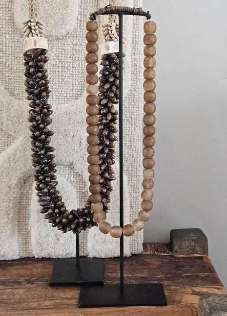 Kralenketting antique lichtbruin naast een donkere schelpenketting op twee zwarte metalen standaarden op een houten ondergrond