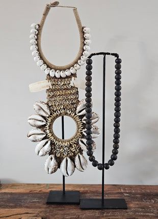 Schelpenketting tijgerschelp op zwarte standaard naast een zwarte kralenketting op standaard op een houten ondergrond voor een witte muur