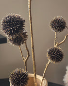 Close-up van bronskleurige Echinops kunsttakken met glanzende stekelige bollen boven een beige vaas tegen grijs