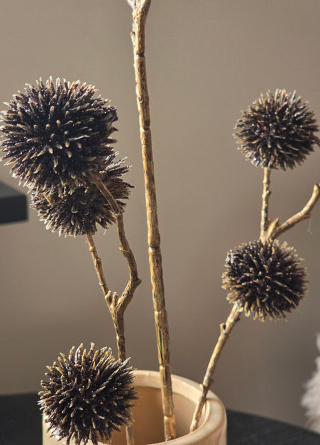 Close-up van bronskleurige Echinops kunsttakken met glanzende stekelige bollen boven een beige vaas tegen grijs