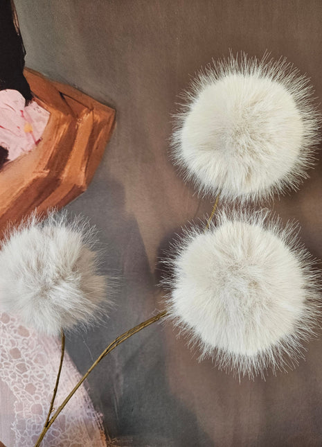 Close-up van drie cream-witte fluffy bollen op dunne bruine stelen op een grijze linnen ondergrond naast een houten dienblad met roze bloemen