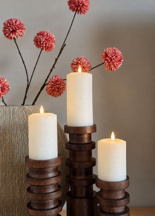 Vooraanzicht van drie cremewitte LED stompkaarsen van wax op houten kandelaars naast een vaas met roze bloemen