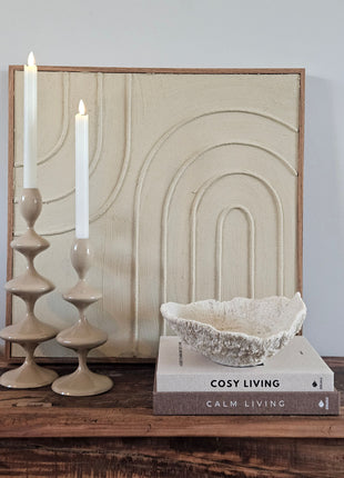 Light & Living ornament schaal Coral crème op koffietafelboeken naast twee taupe keramieke kandelaars voor een crème schilderij met boogmotief op een houten dressoir