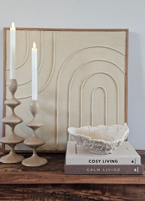 Light & Living ornament schaal Coral crème op koffietafelboeken naast twee taupe keramieke kandelaars voor een crème schilderij met boogmotief op een houten dressoir
