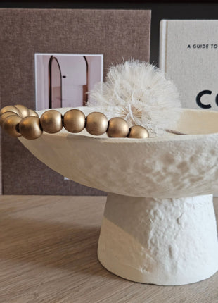 Close-up van Light & Living schaal OTA crème op hoge voet met een gouden kralenketting en witte gedroogde paardenbloem erin voor koffietafelboeken op een houten ondergrond