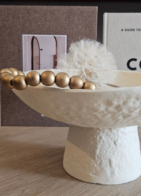Close-up van Light & Living schaal OTA crème op hoge voet met een gouden kralenketting en witte gedroogde paardenbloem erin voor koffietafelboeken op een houten ondergrond