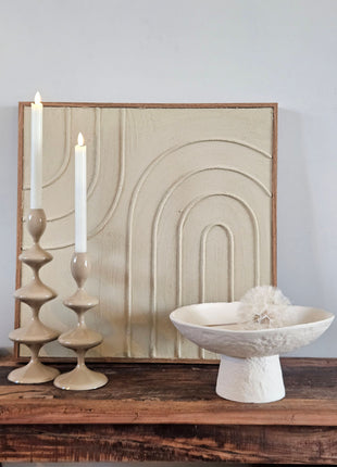 Light & Living schaal OTA crème op hoge voet naast twee taupe keramieke kandelaars voor een crème schilderij met boogmotief op een houten dressoir