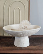 Light & Living schaal OTA crème op hoge voet met ruwe kalksteenstructuur voor een crème schilderij met boogmotief op een houten dressoir met een witte gedroogde paardenbloem erin