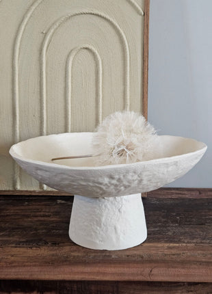 Light & Living schaal OTA crème op hoge voet met ruwe kalksteenstructuur voor een crème schilderij met boogmotief op een houten dressoir met een witte gedroogde paardenbloem erin