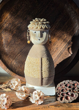 Vooraanzicht van beige Namji Doll Naya met touw en kralen naast droogbloemen en lotus op een houten tafel