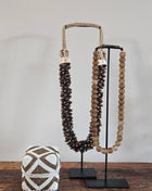 Kleine kralen offermand goud-wit met geometrisch zigzagpatroon op een houten ondergrond naast twee schelpenkettingen op een zwart metalen standaard