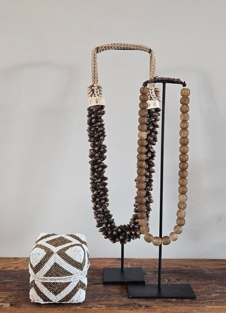 Kleine kralen offermand goud-wit met geometrisch zigzagpatroon op een houten ondergrond naast twee schelpenkettingen op een zwart metalen standaard