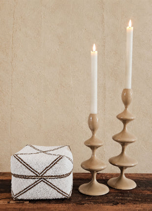 Grote kralen offermand wit-goud met geometrisch kruispatroon op een houten ondergrond naast twee beige keramieken kandelaren met witte kaarsen