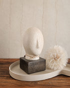 Abstract hoofd beige met ovale vorm en uitstekende neus op een donkerbruine houten voet op een crème dienblad naast een witte gedroogde paardenbloem op een houten ondergrond