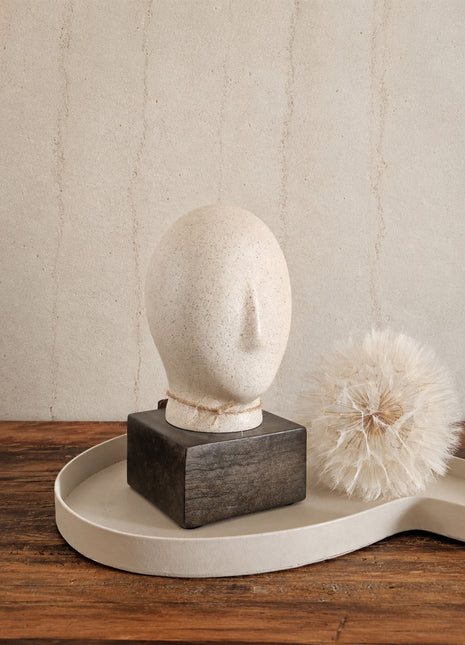 Abstract hoofd beige met ovale vorm en uitstekende neus op een donkerbruine houten voet op een crème dienblad naast een witte gedroogde paardenbloem op een houten ondergrond