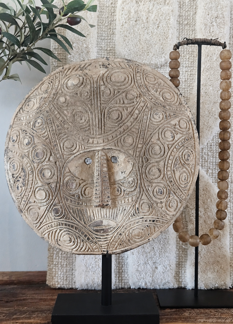 Rond houten masker naturel met gegraveerde gezichtsstructuur op een zwarte metalen standaard naast een houten kralenketting